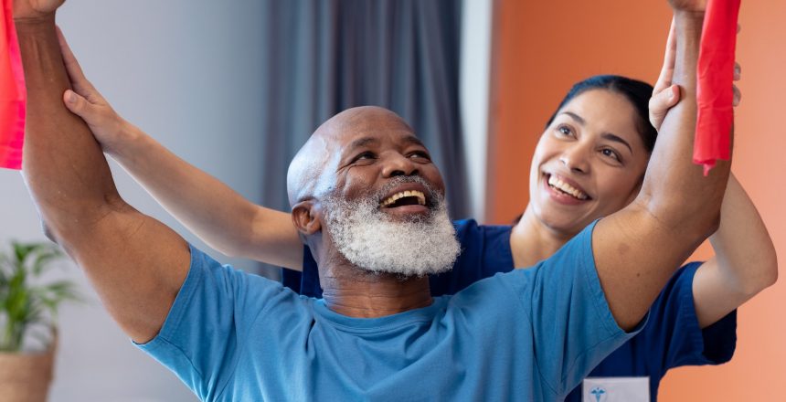 Smiling diverse female physiotherapist and senior male patient exercising with band. Hospital, medical and healthcare services.